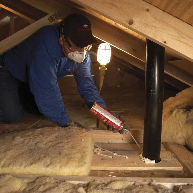 Technician applying spray foam sealant around pipe penetration in attic