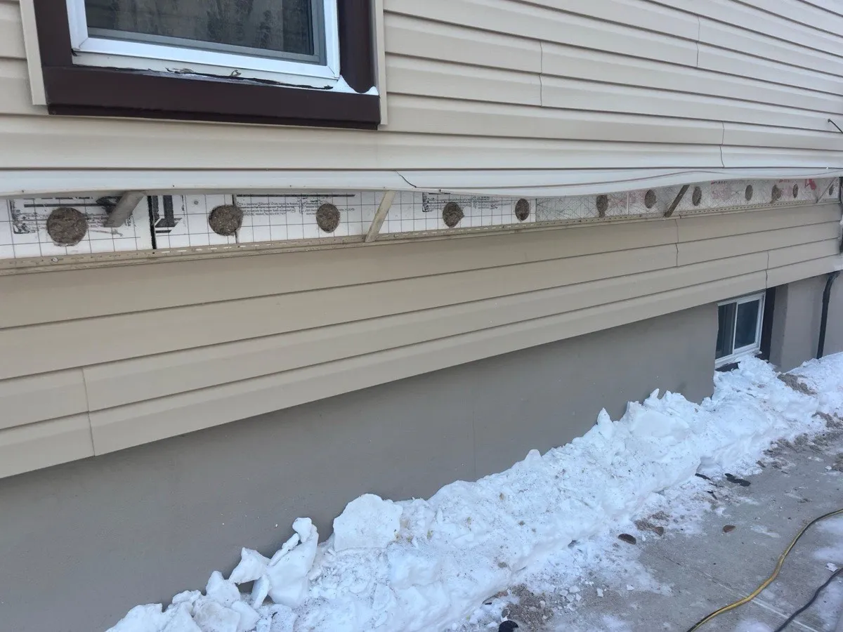 Tan vinyl siding removed showing cellulose-packed drill holes in wall cavity during insulation installation - NY Energy Project